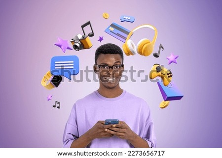 Portrait of cheerful young African American man using smartphone standing near purple wall with headphones, joypad and social media icons. Concept of online entertainment Royalty-Free Stock Photo #2273656717
