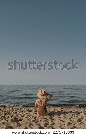 Young woman sitting and relaxing on the beach sand with view on sea. Harmony, lounge, summer vacation concept. Elegant fashion composition with straw hat and bikini swimsuit