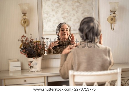 An elderly smiling woman of 80+ years of age spends a good time at home, a grandmother takes care of her appearance, looks in the mirror, sits at a dressing table in her bedroom. Royalty-Free Stock Photo #2273713775