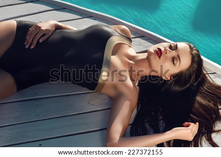 Beautiful brunette girl in good shape with long dark hair and tan skin red lips in black swimming suite with golden line with modern rings lie near the pool with green water closing her eyes and smile