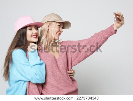 Two young female friends in hipster outfit make selfie on a phone over grey background.