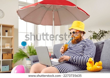 Funny man in classic bathing suit enjoying summer vacation at home. Eccentric bearded man in sunglasses and panama hat sitting under umbrella, drinking cocktail and using laptop in living room Royalty-Free Stock Photo #2273735635