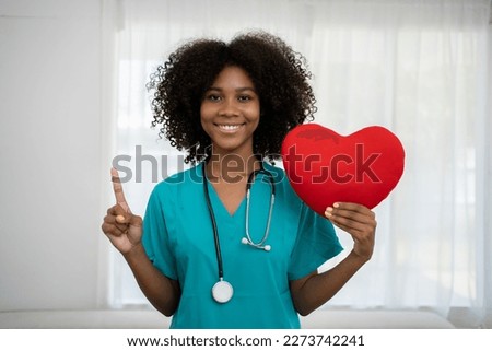 Happy confident African ethnicity female young doctor cardiologist holding heart figure in hands, reminding regular checkup procedures for patients, promoting charity donation or healthcare services. Royalty-Free Stock Photo #2273742241