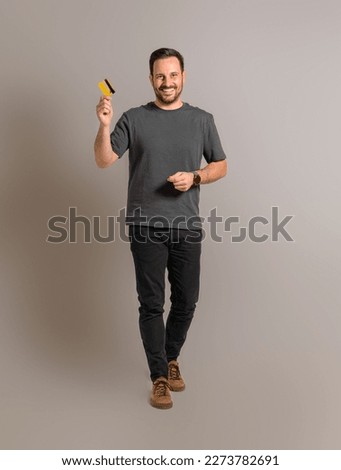 Full length portrait of smiling handsome mid adult man in casual clothing showing credit card while walking isolated against gray background