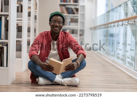 Clever black student man with book sitting on floor in university library. African American guy smiling happily looking at camera, enjoy interesting effective studies in higher institution. Education 
