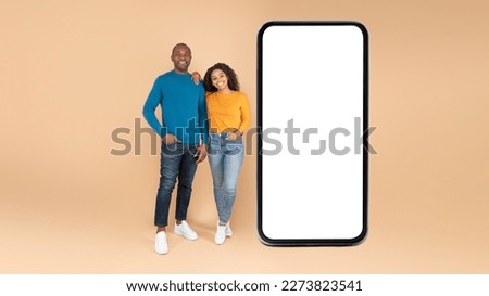 Digital ad and offer. Happy black spouses standing near big cellphone with blank screen, advertising app or website, posing over peach studio background, full length, mockup