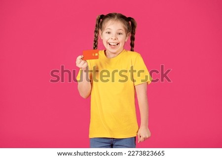Cheerful Cute Little Girl Holding Credit Card In Hand And Looking At Camera, Joyful Excited Preteen Female Child Recommending Bank Services While Standing Isolated Over Pink Background, Copy Space