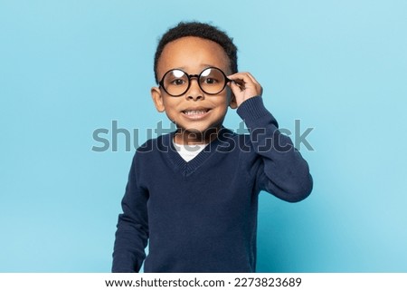 Nerdy african american boy in school uniform touching eyeglasses, looking and smiling at camera, adjusting round glasses against blue studio background