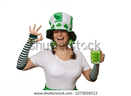 Beautiful young woman with two funny pigtails, wearing Leprechaun carnival hat and clover leaves earrings, showing Ok sign, winking at camera, posing with a mug of green Irish beer on white background
