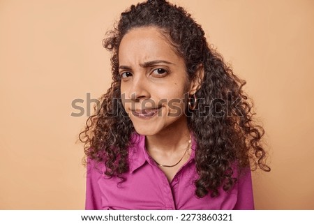 Portrait of young mixed race woman with Afro curly hair looks suspiciously at camera has sarcastic exression listens something that annoys her wearing earrings and pink shirt isolated over brown wall Royalty-Free Stock Photo #2273860321