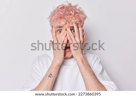 Frightened hipster guy hids face with hands peeks through fingers gasps from fear tries not to see anything has small tattoo on arm dressed in casual t shirt isolated on white background. Omg concept Royalty-Free Stock Photo #2273860395