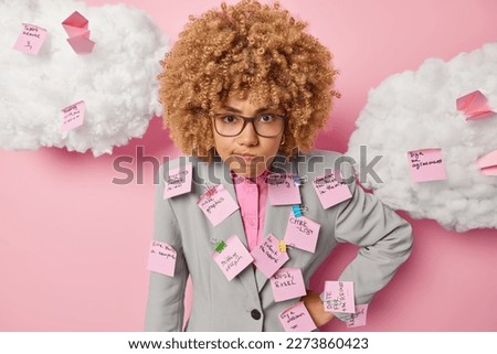 Horizontal shot of displeased annoyed curly haired woman entrepreneur purses lips dressed in formal outfit uses sticky notes for writing down necessary things to do isolated over pink background Royalty-Free Stock Photo #2273860423