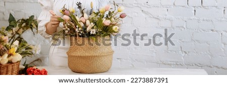 A young woman florist in white clothes and a straw hat stands with a basket of flowers against a white brick wall, in a flower shop. Banner for website header design with copy space. Royalty-Free Stock Photo #2273887391