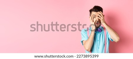 Scared and embarrassed nerdy guy in bow-tie covering his eyes, peeking through fingers at something scary, standing on pink background.