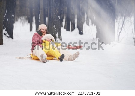 A mother woman with a toddler baby rides an ice sled in a snowy park. Mom and son boy ride in winter from a snow slide. Kid aged one year eight months