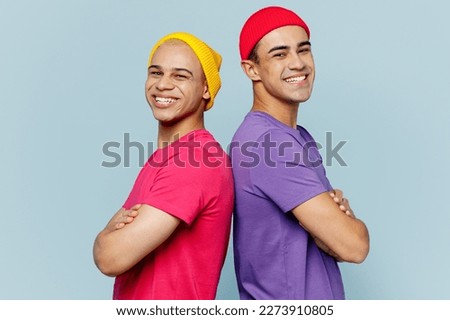 Side view young smiling happy couple two friends men wearing casual clothes looking camera together standing back to back hold hands crossed folded isolated on pastel plain light blue cyan background