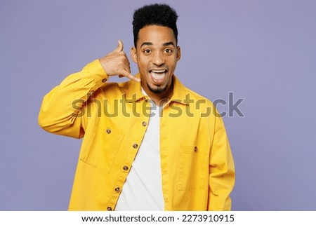 Young man of African American ethnicity wear yellow shirt t-shirt doing phone gesture like says call me back isolated on plain pastel light purple background studio portrait. People lifestyle concept