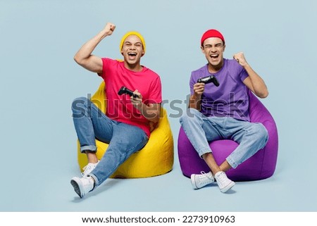 Full body fun young couple two friend men wear casual clothes together sit in bag chair hold play pc game with joystick console do winner gesture isolated on pastel plain light blue cyan background