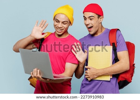 Young IT students couple two friends men wear casual clothes backpack bag together hold books use laptop pc comouter waving hand isolated on plain pastel blue background. High school college concept