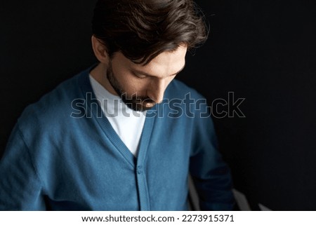 Sad upset bearded handsome young man having troubles at work, failed in business project or lost big sum of money investing at stock trading market, looking down against black background