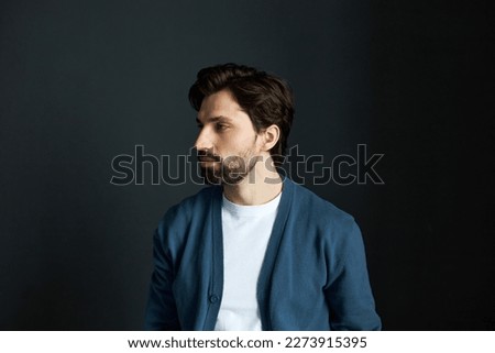 Portrait of handsome millennial male math teacher with beard in blue cardigan posing against black studio background turning head to the right, thinking of educational plan for week