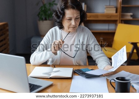 Middle aged senior woman sit with laptop and paper document. Pensive older mature lady reading paper bill pay online at home managing bank finances calculating taxes planning loan debt pension payment Royalty-Free Stock Photo #2273937181
