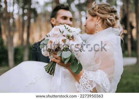 A stylish groom in a suit holds in his arms a beautiful bride in a long white dress, with a bouquet of roses, flowers in a park, forest. Close-up wedding photography, portrait of smiling, newlyweds.