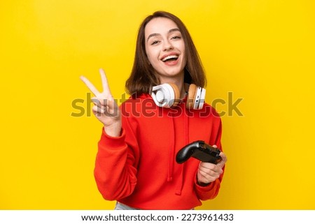 Young Ukrainian woman playing with a video game controller isolated on yellow background smiling and showing victory sign
