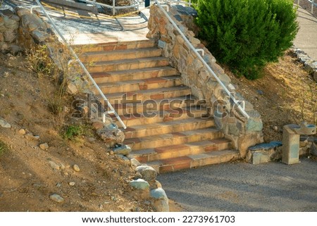 Cement stone and rock stairs steps with metal hand rail and asphalt with dry grass and shrub nearby. In late afternoon shade in suburban neighborhood near house or home in park or walkway.