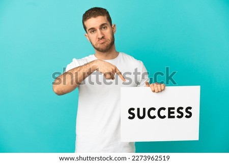 Handsome blonde man over isolated blue background holding a placard with text SUCCESS and  pointing it