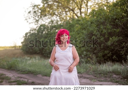 Bright diversity woman with pink hair portrait. The girl squatted down on a forest path into the sunset. Childfree girl, weekend trip. Individual, bold style. Green nature background. Copy space