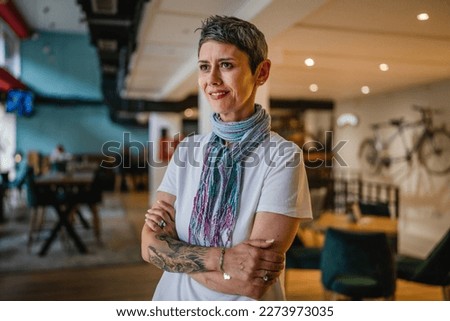 One woman senior caucasian female with short gray hair modern look stand in cafe or restaurant alone wear white shirt tattoo on hand confident happy[y smile real person copy space Royalty-Free Stock Photo #2273973035