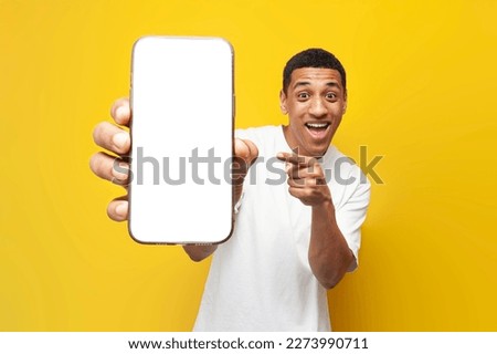 young african american guy in white t-shirt shows blank screen of smartphone on yellow isolated background, man holds phone and advertises screen for mock-up Royalty-Free Stock Photo #2273990711