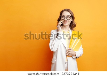 young business girl in glasses and white suit holds documents and speaks on the phone on colored isolated background, female student in business clothes communicates on mobile phone