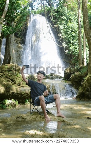 asian man sitting relaxed while making a video call against the backdrop of the beautiful Randusari waterfall in Bantul, Yogyakarta