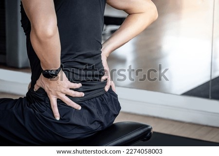 Sport man feeling lower back pain or spine pain while sitting on workout bench in fitness gym. Male athlete suffering from sport injuries symptoms. Royalty-Free Stock Photo #2274058003