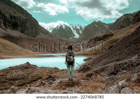 A tourist girl with a large backpack stands near an alpine lake in the stones in the Altai mountains against the backdrop of glaciers and snow in autumn. Royalty-Free Stock Photo #2274068785
