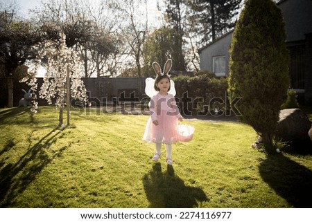 Little girl in pink fairy dress with wings and bunny ears holding pink basket looking around during an Easter Egg hunt in a front yard in Edinburgh, Scotland, UK, during a sunny day in Easter