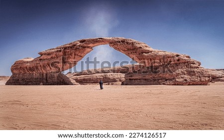 A "Dust Devil" or small tornado passes through "Rainbow Rock", also known as "Arch Rock' in Alula, Saudi Arabia. A man wearing traditional Clothes stands right under the arch.  Royalty-Free Stock Photo #2274126517