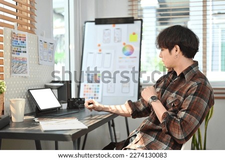 Young asian male web developer in plaid shirt using digital tablet and working with layout app design project at office
