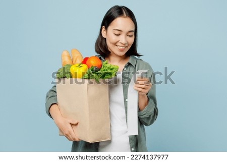 Young smiling happy fun woman wear casual clothes hold brown paper bag with food products look at check isolated on plain blue cyan background studio portrait. Delivery service from shop or restaurant Royalty-Free Stock Photo #2274137977