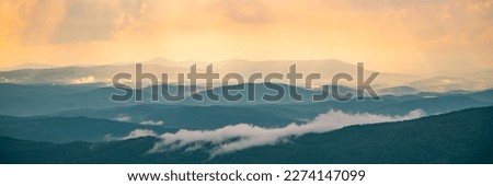 Panorama of the mountains at sunset over a dark mountain. Colorful sunrise with clouds over the hill. The sun hides behind a cloud in the daytime sky. Royalty-Free Stock Photo #2274147099
