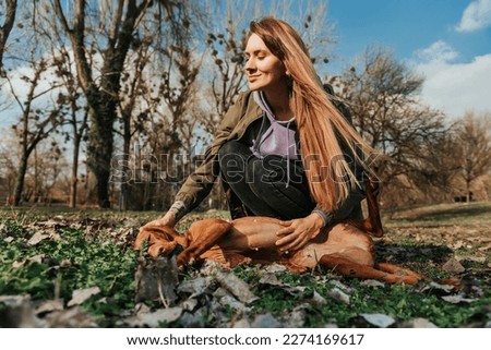 Young woman petting dog while lying on ground in park. Hungarian vizsla with ball in mouth lying in grass getting belly rubs from girl in casual outfit. Happy female with dog enjoying walk outdoors. Royalty-Free Stock Photo #2274169617