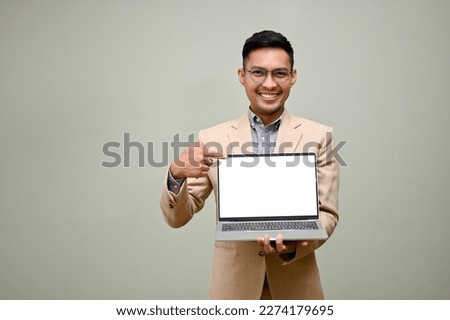 Happy and smiling adult Asian businessman in formal business suit holding his laptop, showing laptop blank screen, pointing his finger at laptop screen, standing against green background.