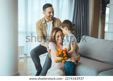 Cheerful little boy with card and with bouquet of tulip flowers smiling and congratulating happy mom on mother day at home. Woman looking at adorable son while sitting on floor with mothers day card