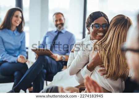Caring female counselor hugs a female patient during a group therapy session. A young mixed race woman hugs a mature adult woman. They are sitting next to each other in a medical clinic. 