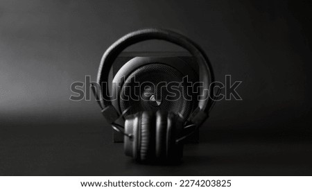 Black one-way speaker is visible through black headphones with ear pads on a black background. Concept of modern technology and wireless audio. Copy space. Selective focus. Defocus. Close-up