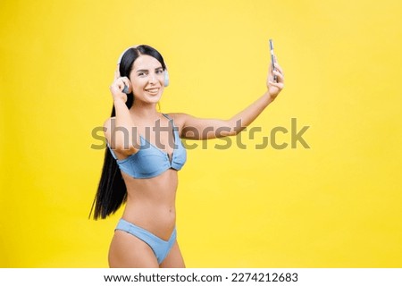Portrait of young pretty girl with wireless headphones on head, taking selfie for social networks, smiling and laughing at phone camera, isolated on yellow background.