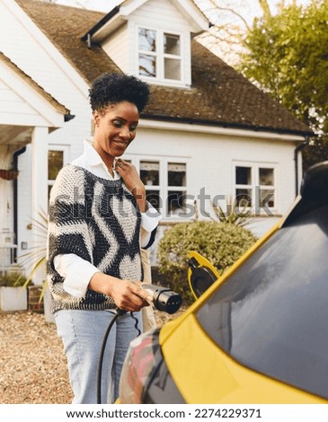 Woman charging electric car on driveway outside house Royalty-Free Stock Photo #2274229371