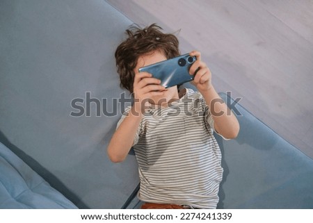 portrays a child engrossed in his phone, sparking a debate on the impact of technology on young minds and how parents can monitor and limit screen time. Royalty-Free Stock Photo #2274241339